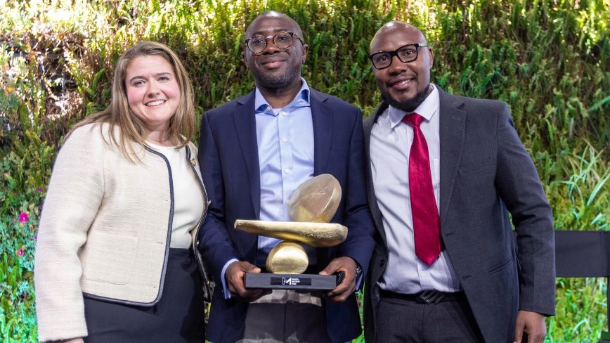Three individuals stand together smiling in front of a lush green backdrop. The person in the center holds a golden sculptural trophy with a base labeled “Milken-Motsepe Prize.” This image depicts the award presentation for the Milken-Motsepe Prize in FinTech, celebrating innovation in financial technology.