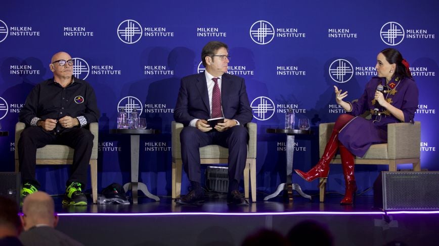 Three panelists seated onstage at the Milken Institute Global Investors' Symposium in Mexico City 2025. The panel includes a man in neon sneakers and black shirt, a man in a business suit holding a notebook, and a woman in a purple dress and red boots speaking animatedly. The Milken Institute logo is displayed on the backdrop.
