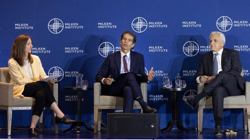 Three professionals seated onstage during a panel discussion at the Milken Institute Global Investors' Symposium Mexico City 2025. A woman in a cream blazer, a man in a dark suit gesturing while speaking, and another suited man listen attentively. The Milken Institute logo appears repeatedly on the blue backdrop.