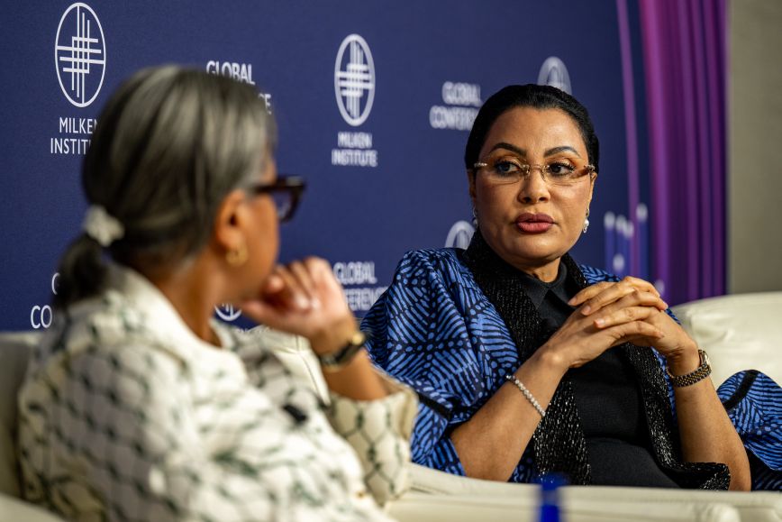 Dr. Doris Uzoka-Anite, wearing glasses and a blue patterned jacket, listens intently with hands clasped while speaking on stage to another panelist in a white and green patterned outfit, against a “Milken Institute Global Conference” backdrop.