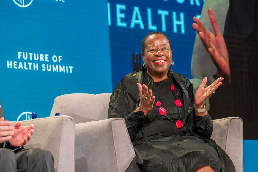 A smiling speaker applauds on stage during a session at the Milken Institute’s Future of Health Summit. She is seated in a gray armchair, wearing a black textured dress with bold red jewelry, set against a blue backdrop featuring the event's name, “Future of Health Summit.”