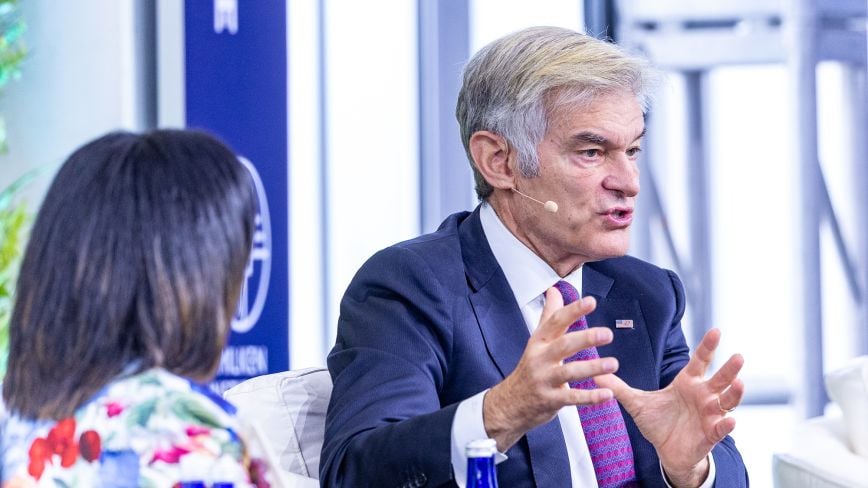 A man identified as Mehmet Oz, wearing a navy suit, white shirt, and patterned pink tie, speaks animatedly into a headset microphone, gesturing with both hands. He is seated on stage in conversation with a woman in a colorful floral blouse, whose back is to the camera. The background shows a blurred event setting with signage.