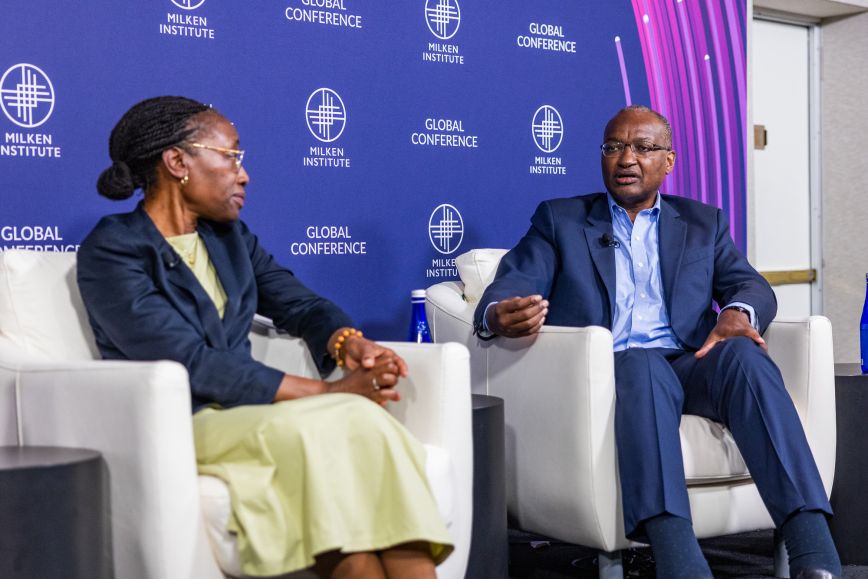 Two panelists seated in white chairs, speaking on stage in front of a dark blue “Milken Institute Global Conference” backdrop—one wearing a dark blazer and light green dress, the other in a navy suit, gesturing while responding to the discussion.