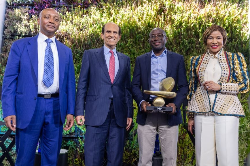 Four individuals standing together on stage, smiling at the camera in front of a green foliage wall; one man holds a large gold award sculpture. Dr. Precious Moloi-Motsepe stands at the far right wearing a patterned jacket and white trousers.