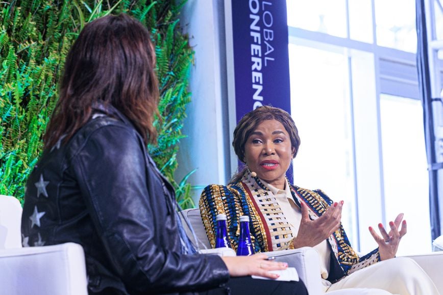 Dr. Precious Moloi-Motsepe, wearing a patterned jacket over a white blouse, speaks with expressive hand gestures during a panel discussion, seated across from another speaker in a black leather jacket with white stars, against a backdrop of greenery and a “Global Conference” banner.