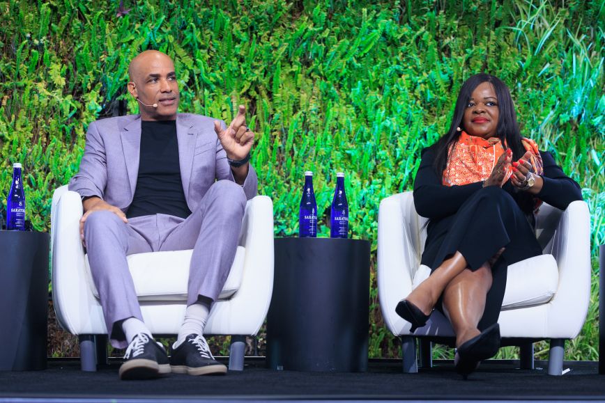 A man in a light purple suit and black shirt speaks while gesturing with his hand, seated next to a woman in a black outfit with an orange patterned scarf, both participating in a panel discussion on stage with a lush green plant wall backdrop.
