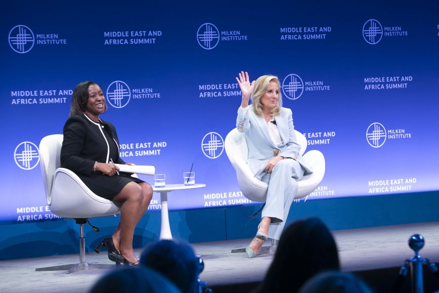 Two women sit on stage at the Milken Institute Middle East and Africa Summit. One is wearing a black dress with white trim and smiling, while the other, dressed in a light blue pantsuit, waves to the audience. Both are seated in modern white chairs with a small table and water glasses between them.