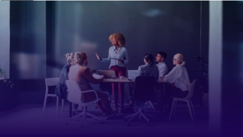 A diverse group of professionals in a business meeting, with a woman standing and leading a presentation at the head of a conference table.