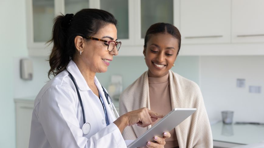 A female doctor in a white coat and stethoscope shows information on a tablet to a smiling female patient during a medical consultation.
