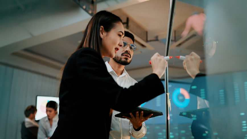 Two business professionals collaborate at a glass board, with one writing on it with a marker while the other holds a tablet.