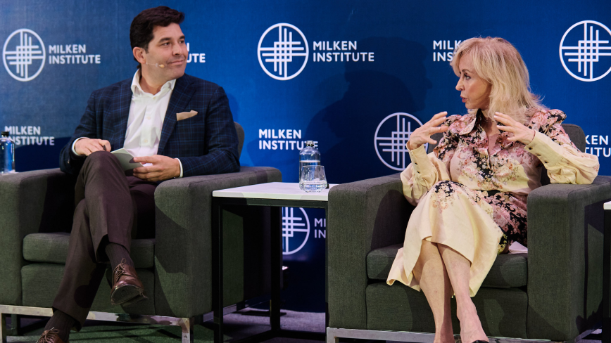 Two people sitting in armchairs on stage during a panel discussion with blue branded backdrop