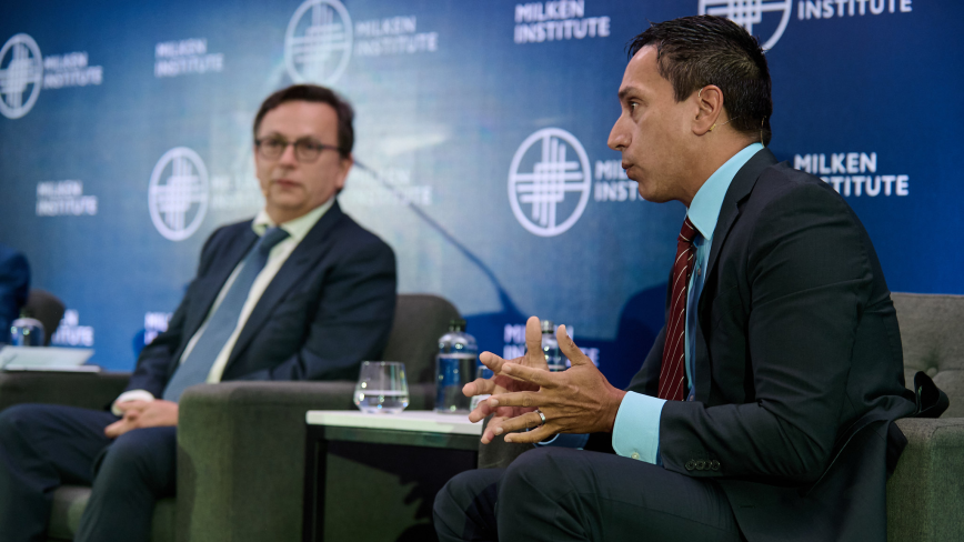 Two men in business suits engaged in conversation at a panel discussion with branded backdrop