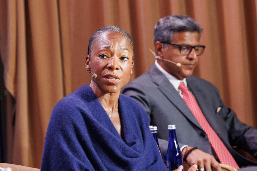 Two professionals seated at a panel, woman in blue suit speaking into microphone, man in glasses beside her