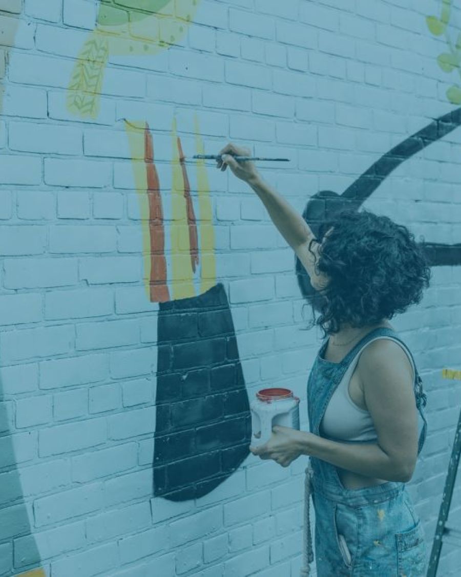 An image with a blue-ish overlay of a person wearing overalls with short black curly hair, painting a mural on a brick wall.