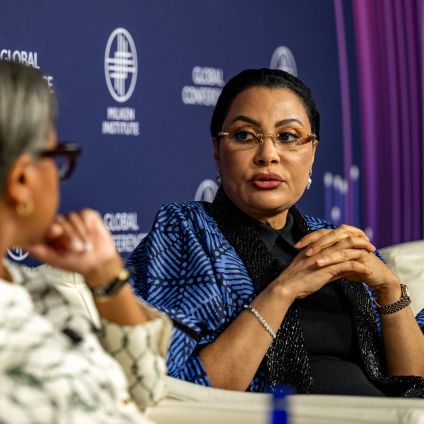 A panelist listens attentively during a discussion at the Milken Institute Global Conference, with a branded backdrop.