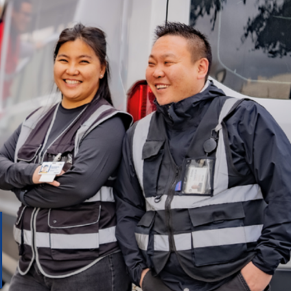 Two smiling employees in reflective safety vests and ID badges lean against a white transport van.