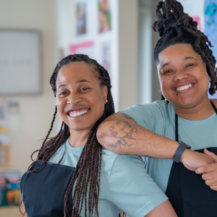 Two smiling women in light blue shirts and black aprons pose closely together in a brightly decorated room featuring children's artwork.