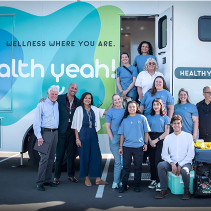 A diverse group of healthcare staff and professionals pose in front of a large "Health Yeah!" mobile wellness clinic parked outdoors.