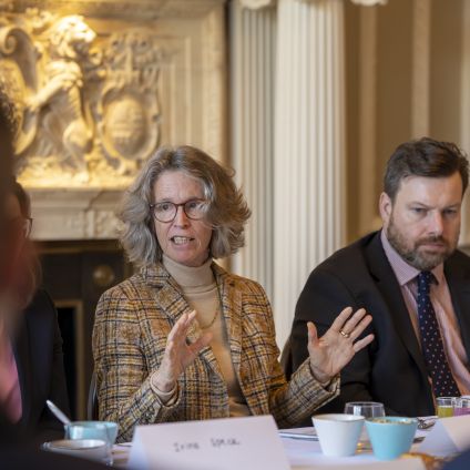 A woman with gray hair and glasses, wearing a plaid blazer, speaks animatedly with both hands raised while seated at a formal conference table. A man in a suit and tie sits attentively beside her. An ornate fireplace with carved stonework is visible in the background.
