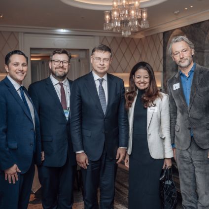 Five people pose together for a photo in an elegantly decorated event venue with a chandelier overhead. The group consists of four men and one woman, dressed in business attire, smiling at the camera.
