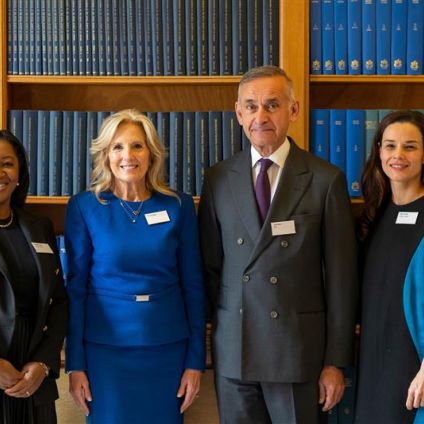 Four people — three women and one man — stand together smiling in front of a wooden bookshelf filled with matching blue bound volumes. All are wearing name badges and dressed in professional attire.