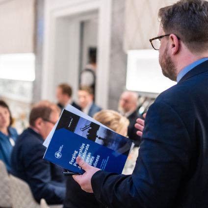 A man in a dark suit and glasses stands with his back to the camera, holding a blue report titled "Forging Partnerships to Accelerate European Readiness," while addressing a seated audience at a professional event.