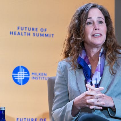 Woman with curly hair speaking at podium wearing gray blazer against yellow backdrop with blue logos