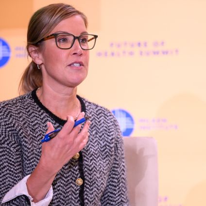 Woman with glasses speaking into a microphone at an event with branded backdrop
