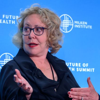 Woman with glasses speaking at World Health Summit, hands gesturing, against blue backdrop