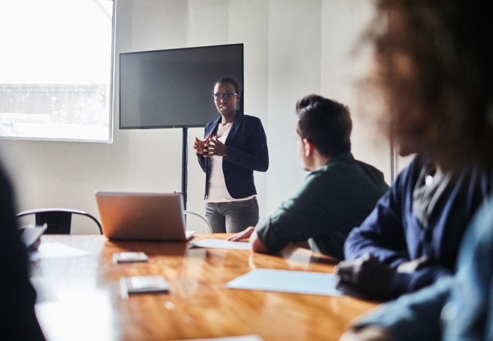 A Black female standing giving a presentation to other people sitting around a conference table