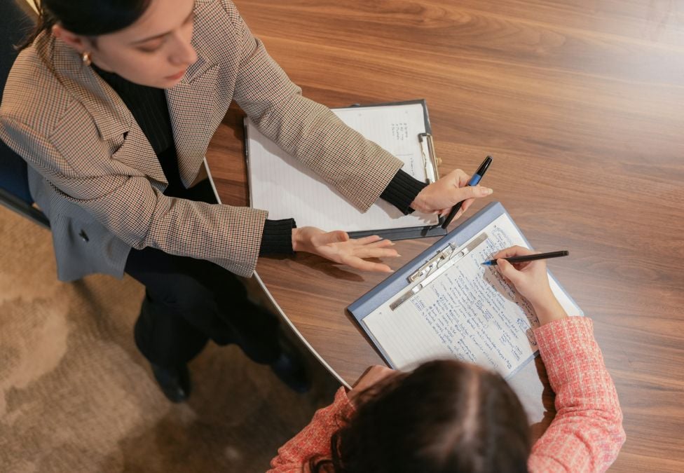 Two female-presenting people sit at a table together going over paperwork