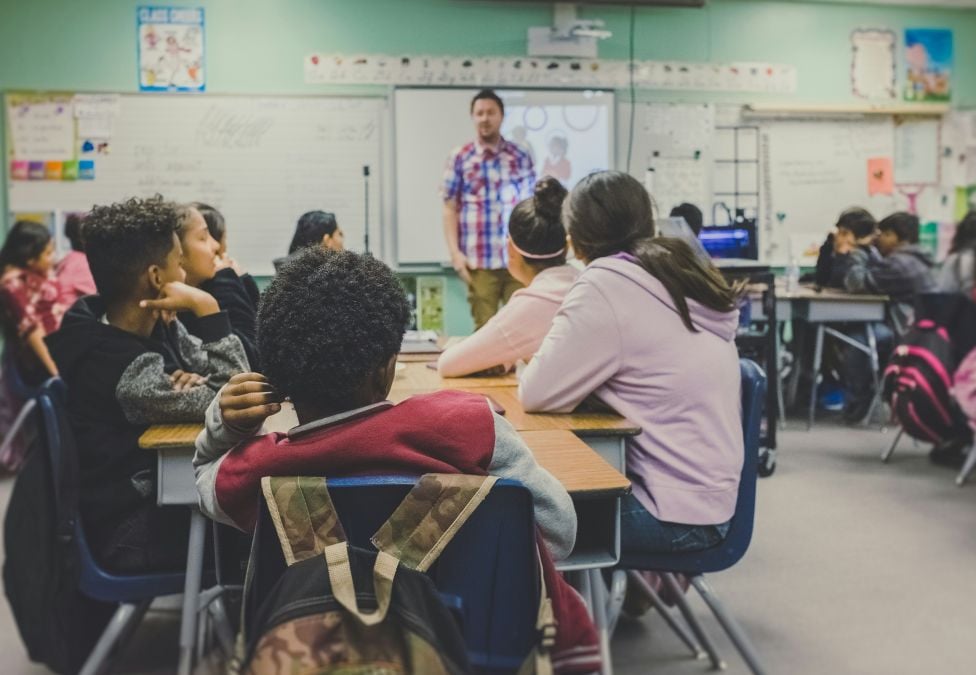 A teacher in front of a classroom of fifth graders sitting around tables in a classroom