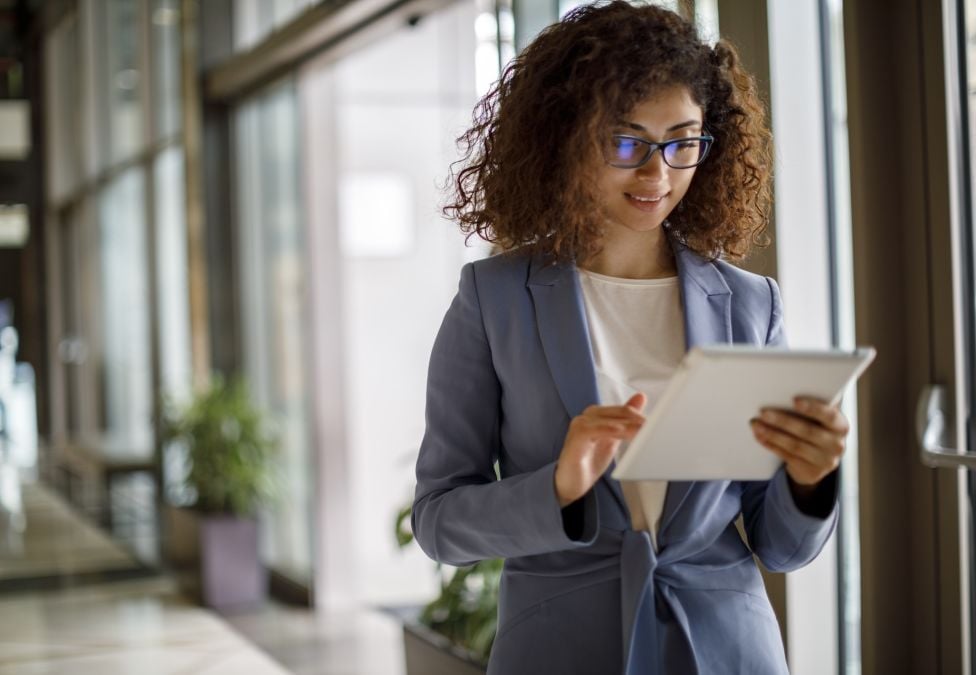 Female-presenting individual with darker skin and brown curly hair wearing a blue suit stands with papers in her hand in an office setting