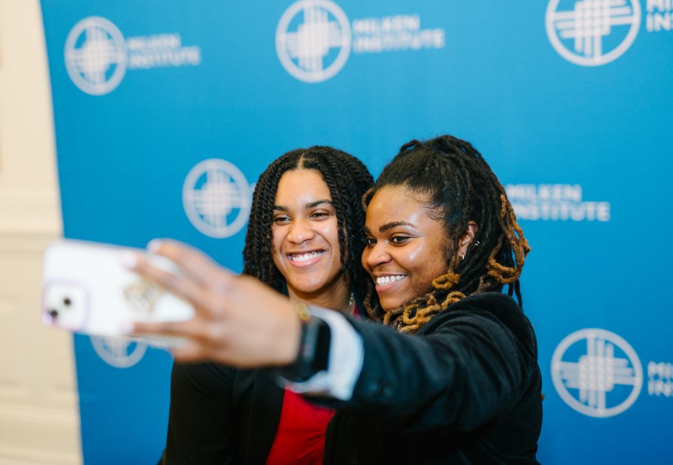 Two young Black women take a selfie in front of a step and repeat backdrop