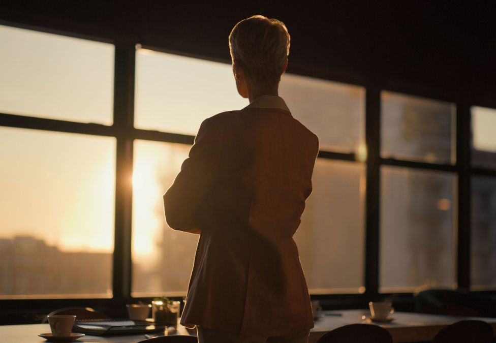 A person, photographed from behind, stands in an office and looks out the window as the sun sets.