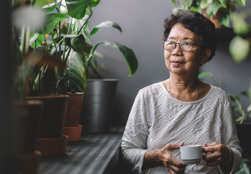 person sitting in room with plants, holding a mug of a beverage staring to their right