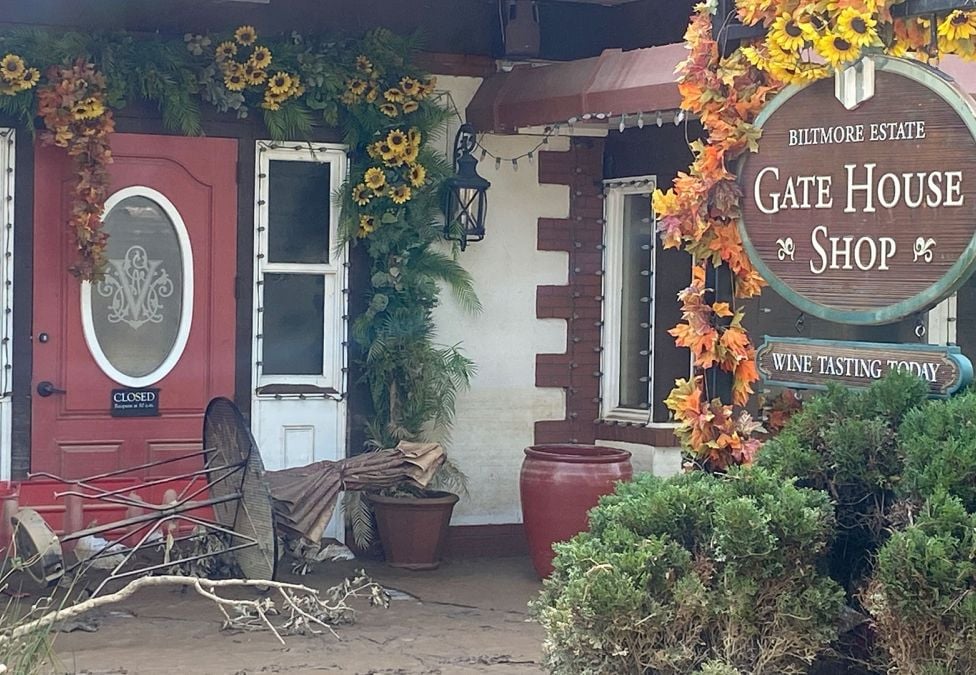 An image of the outside of the Biltmore Estate's Gate House Shop. The building features a red door, surrounded by flowers, and shows a knocked over table and chairs due to the aftermath of hurricane activity.