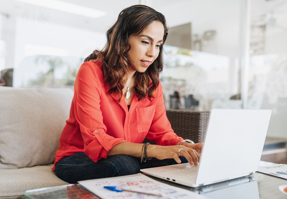 person with dark mid-length hair wearing coral button up blouse sitting on a white sofa, typing on laptop on coffee table, depicting Supporting Family Caregiving report for Milken Institute 