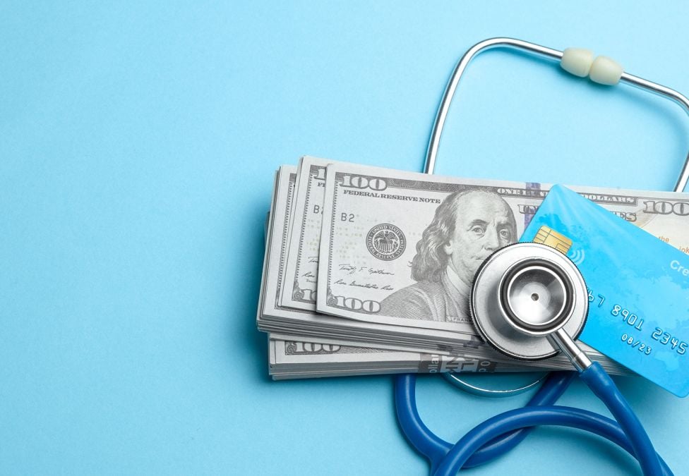 A hero image for an article titled "The Wealth-Health Connection: How Economic Status Shapes Well-Being". It is an image with a light blue background and pictures a stethoscope wrapped around a pile of US dollars and a credit card, signifying wealth and health.