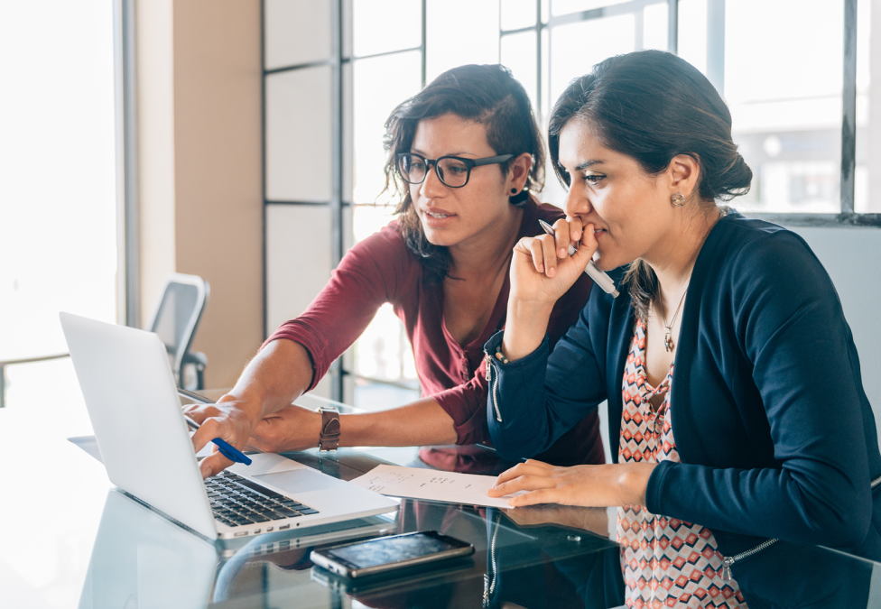 A photo of two Latin American women with dark hair and professional attire sitting in an office and looking at a shared laptop.