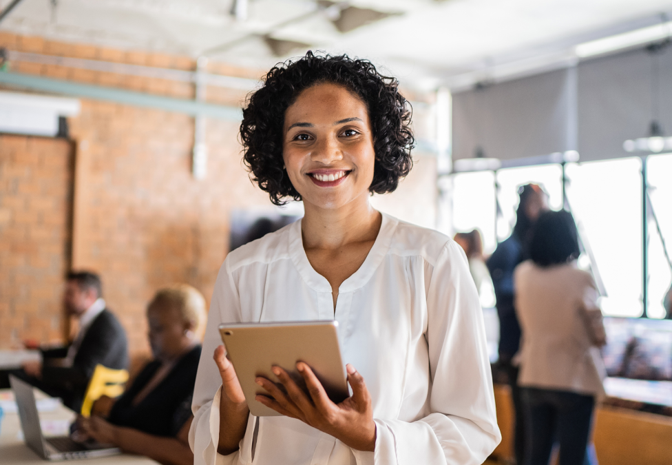 A photo of a woman standing in an office environment, smiling, and holding a tablet. She has tan skin and short dark curly hair, and is wearing a white long-sleeved shirt.