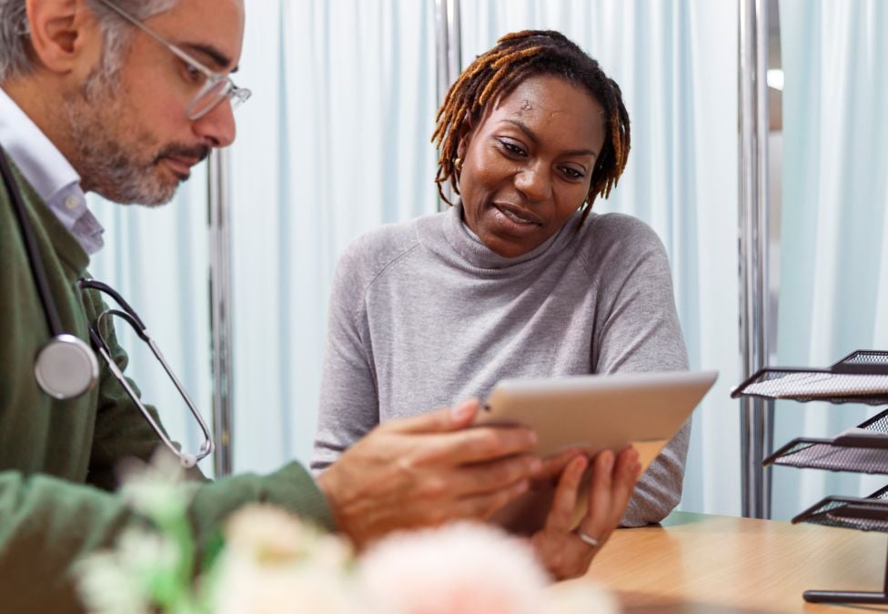 An image of a woman with brown skin and short brown braids, wearing a grey sweater, talking to a male doctor with grey hair, a gray beard, white skin, glasses, a green sweater, and wearing a stethoscope. They are reviewing a tablet together and the image leads to a Milken Institute article around a preventative health roundtable. 