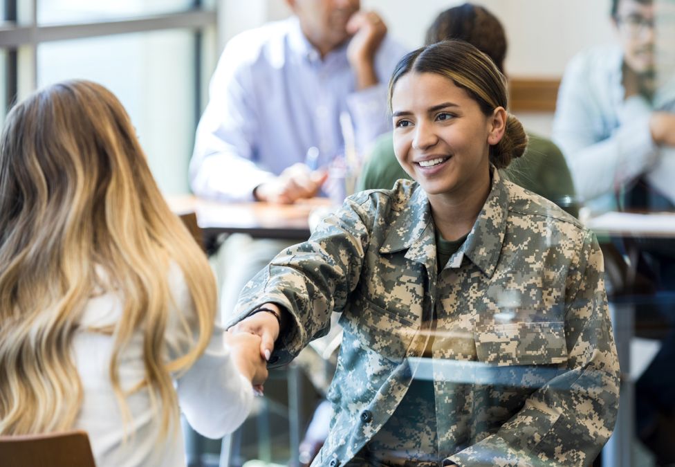 An image of a woman with white skin and brown hair in a bun, wearing a military uniform and shaking hands with a blonde woman with white skin. The image leads into an article about reinvigorating the career pathways for veterans.