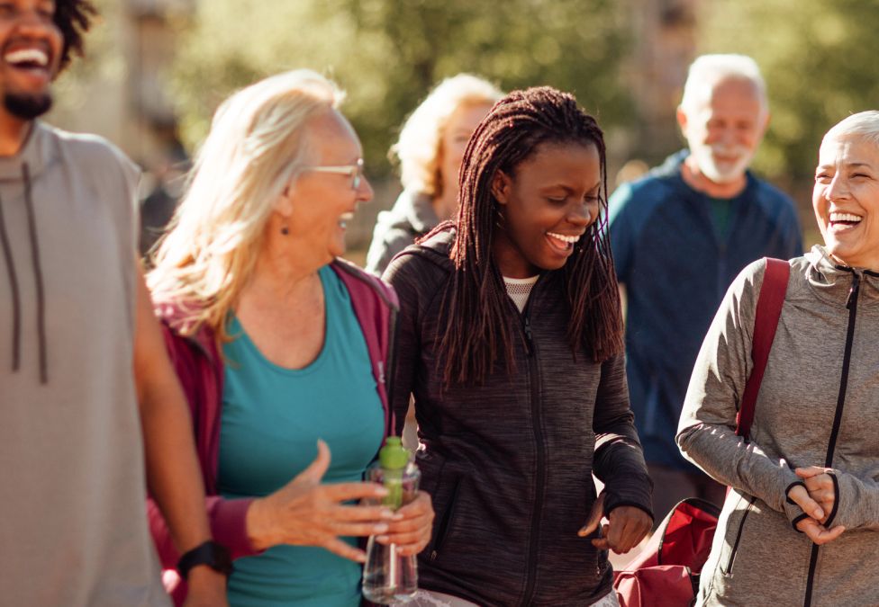 An image of a young man with dark skin, an old woman with white skin, a young woman with dark skin, and another old woman with white skin, standing together and laughing with people and nature in the background. This image leads to an article by the Milken Institute about Scaling and Sustaining Better Health Outcomes Through Prevention.