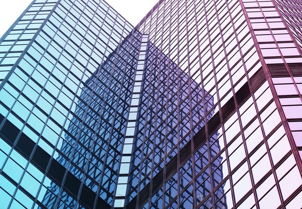 An image of a skyscraper building, upclose, pointing upwards and showing its glass windows with blue-ish and purple-ish hues reflected off of the windows.