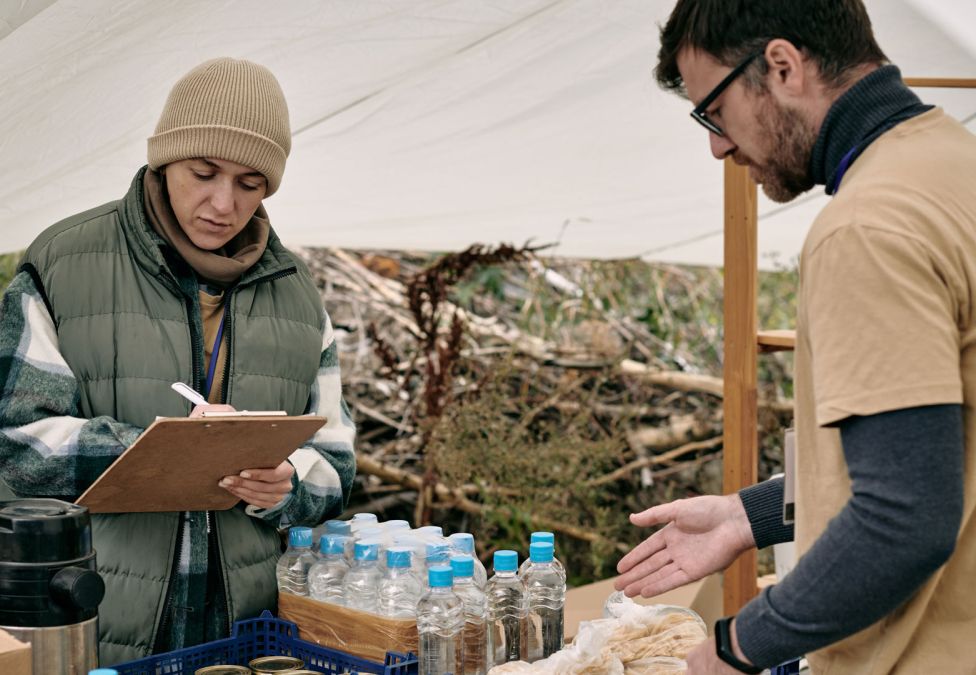 An image of a man and a woman, each with white skin and brown hair, standing under a tent. The woman is checking off a checklist on a clipboard and the man is pointing to a case of water, indicating a disaster relief tent.