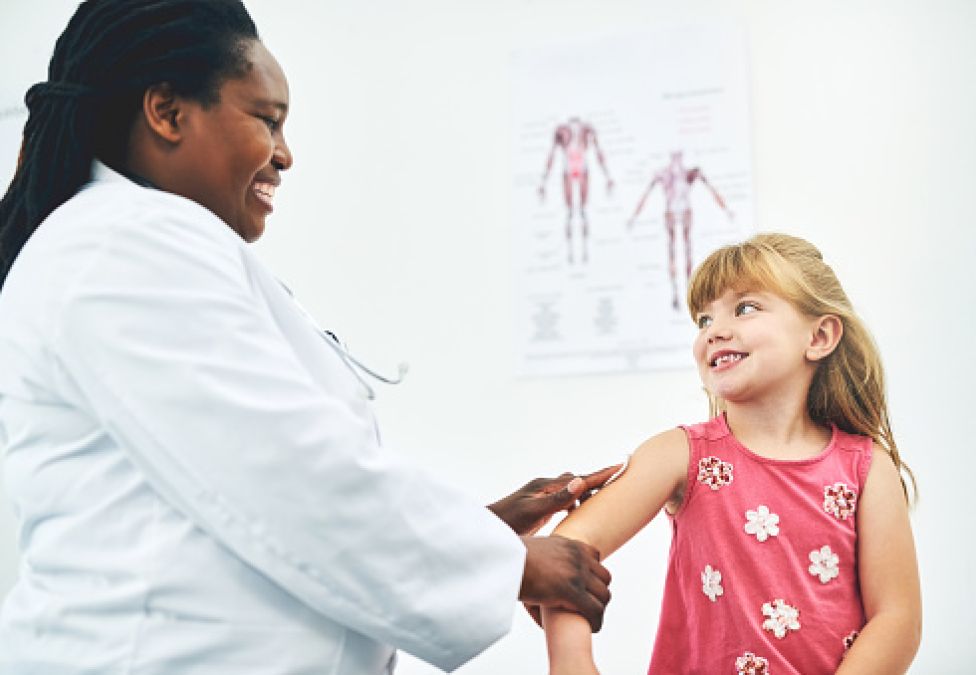 An image of a woman doctor with brown skin and long black hair, wearing a white lab coat, tending to a girl child patient with white skin, blonde hair with bangs, and wearing a pink shirt with flowers on it.