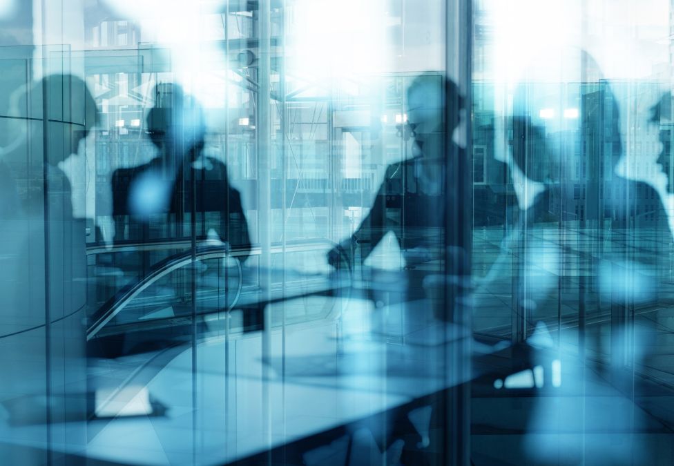 An abstract image with a blue-ish tint of a large window reflection that shows vague figures of business people sitting at a table conducting a meeting.