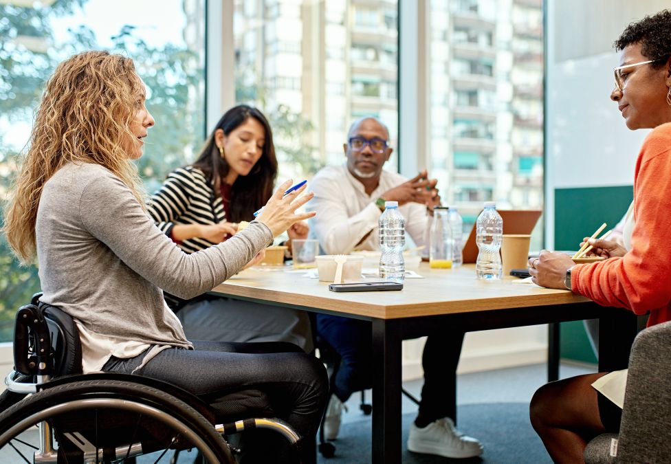 An image of four diverse adults sitting around a table having a discussion in an office space. One appears to be a woman with blonde curly hair, white skin, and a gray sweater sitting in a wheelchair. Another appears to be a woman with brown skin, short brown hair, glasses, and an orange shirt. Another appears to be a woman who is Asian, with white skin and brown hair wearing a striped top. And the other person appears to be a man with brown skin, a bald head, who is wearing business attire.