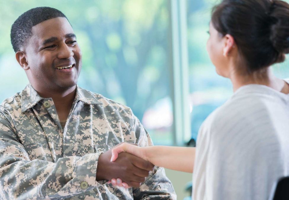An image of a smiling man with brown skin and short brown hair, wearing camouflage military attire, shaking hands with a woman with white skin and brown hair in a bun, wearing a white shirt.